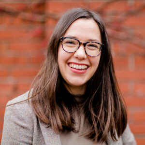 A headshot of Kayla. She is a young white disabled woman with straight brunette hair and glasses. In this photo, she is looking right at the camera, laughing. There is a red brick wall out of focus behind her.