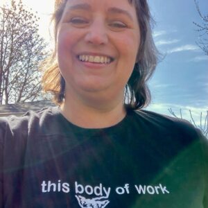 selfie of the artist, a caucasian woman in a black T shirt that reads "this body of work", smiling against a sunny blue sky background..
