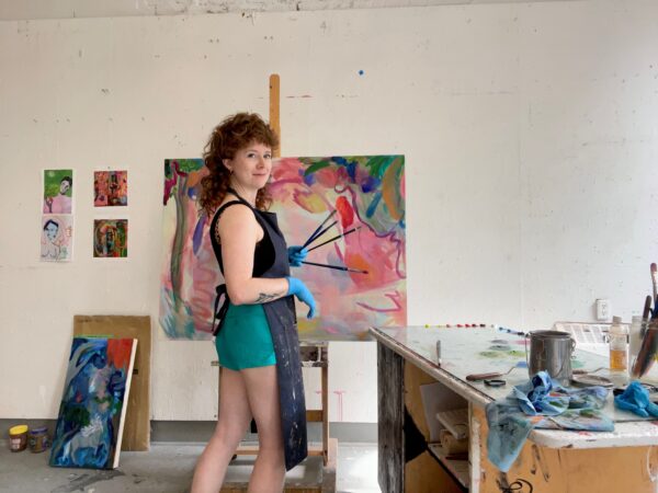 Cortney Cassidy, a white genderqueer person, stands in front of an easel with a large brightly coloured painting with paintbrushes in their hands. They are standing in a painting studio, with various paintings and paint supplies in the background.